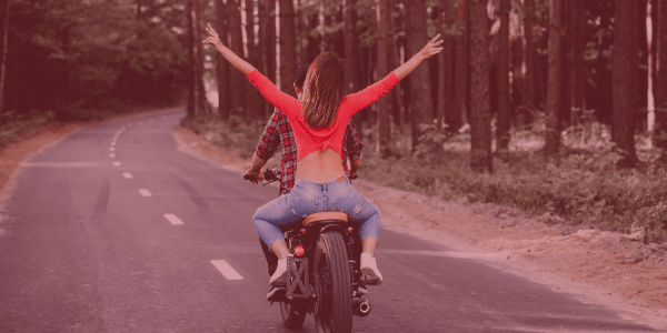 A driver and passenger on a motorcycle on a road in a wooded area with passenger having her hands in the air